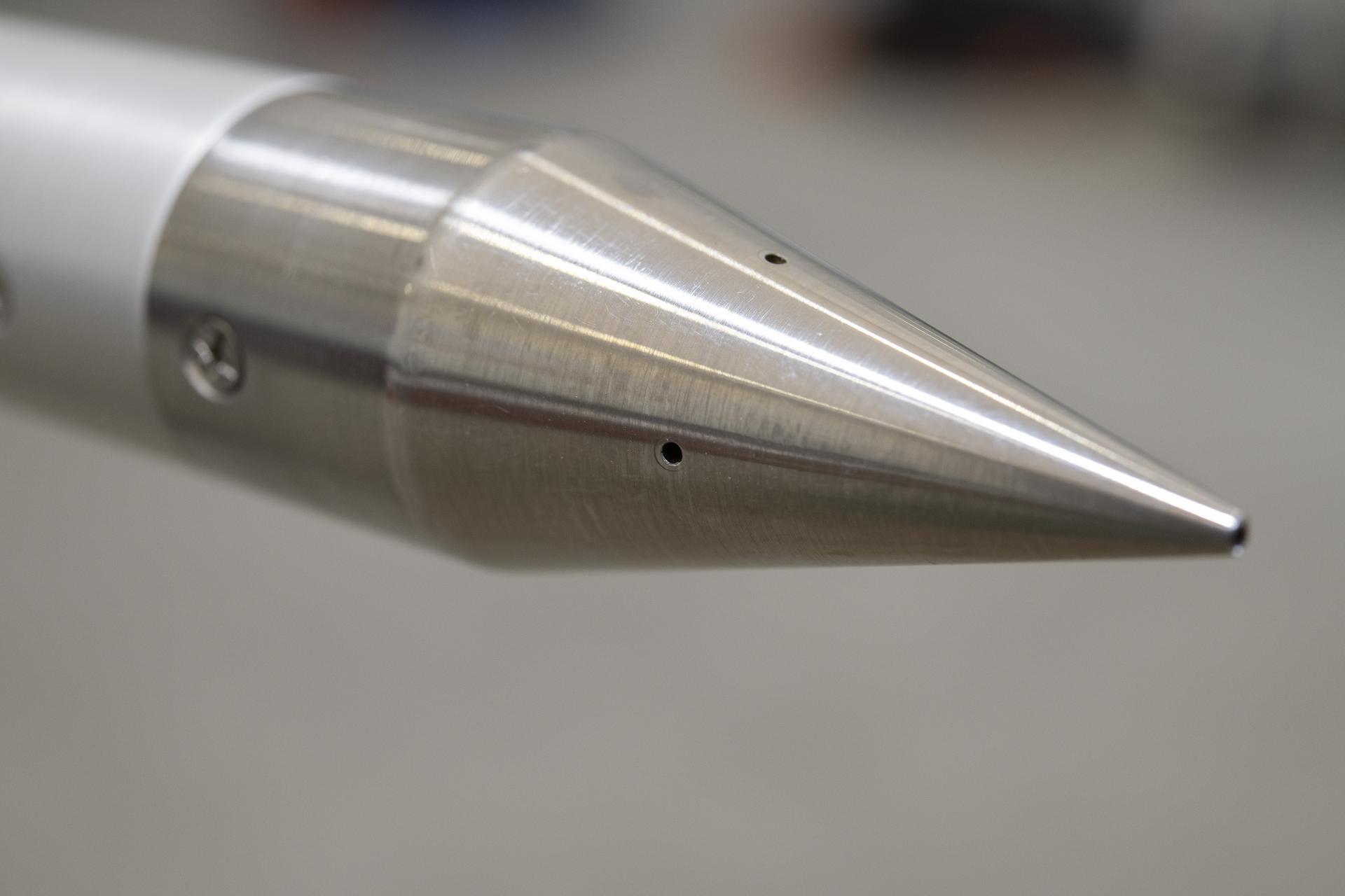 A silver cone is pictured, highlighting small measurement holes that act as pressure ports, evenly spaced at the tip and around the shape of a cone. The probe will used to measure shock waves during supersonic flight tests.