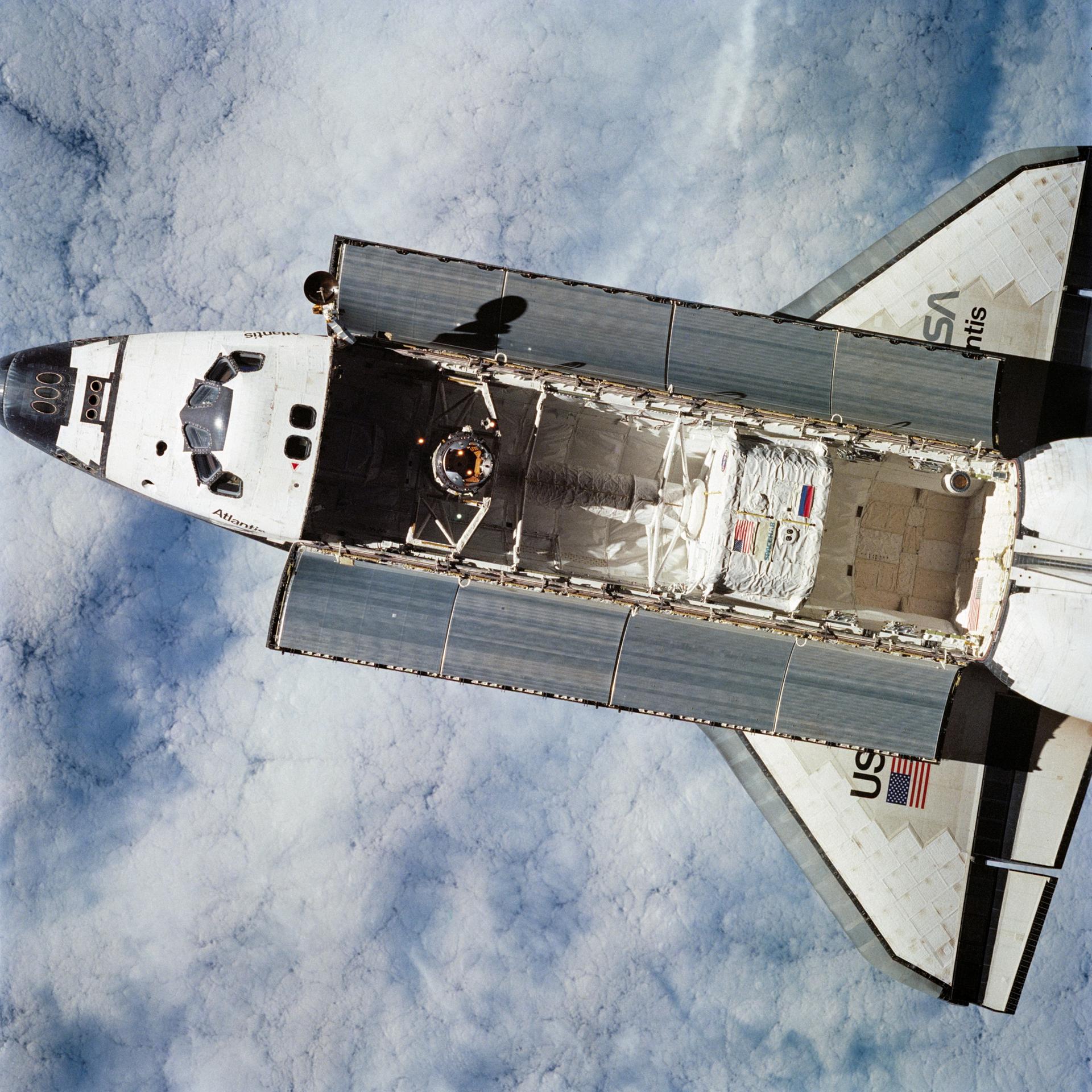 View of Space Shuttle Atlantis from above. It has its payload bay doors open. The Earth, covered in clouds, can be seen below.